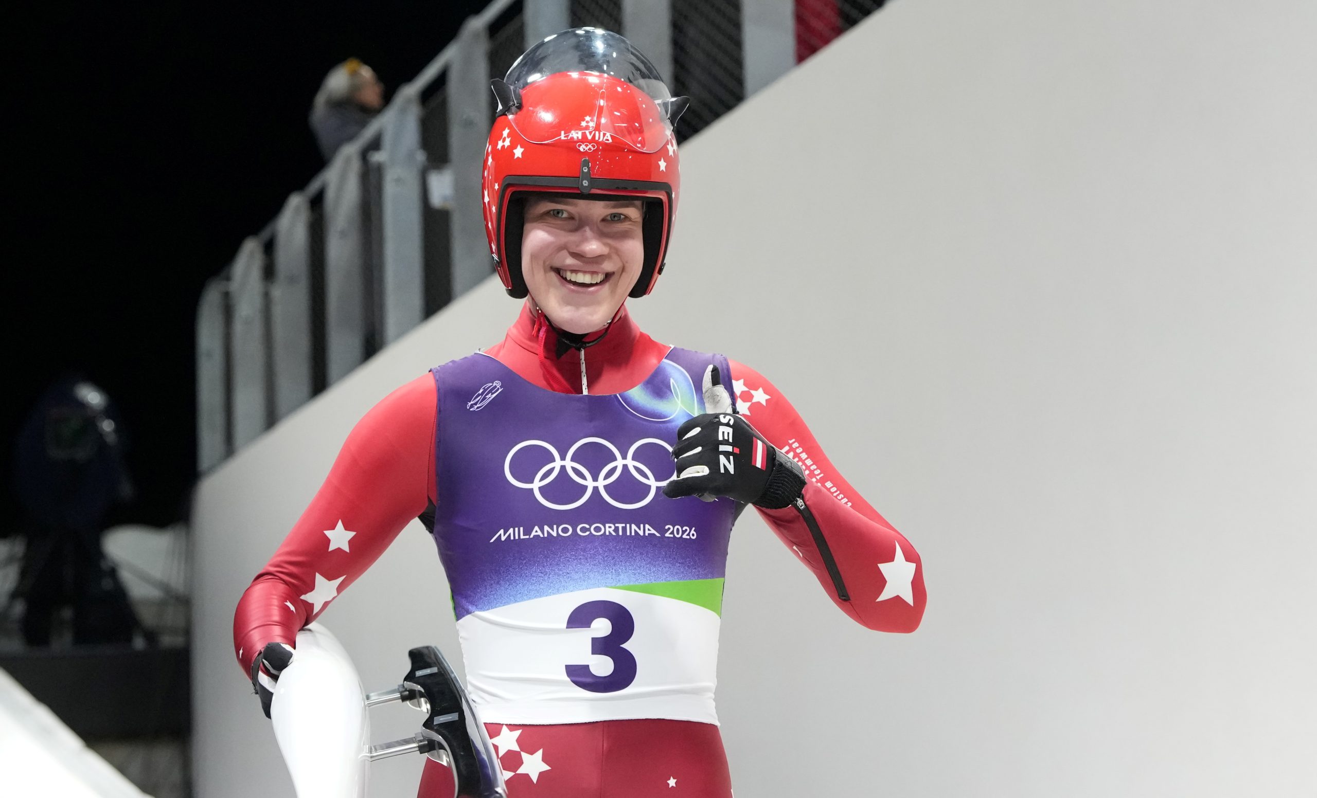 Latvijas kamaniņu sportists ar starta numuru 3 smaida trasē, turot kamanas, gatavojoties braucienam.