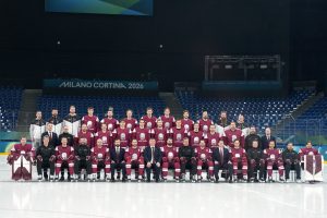 Latvijas hokeja izlase kopbildē uz ledus arēnā ar uzrakstu “Milano Cortina 2026”.