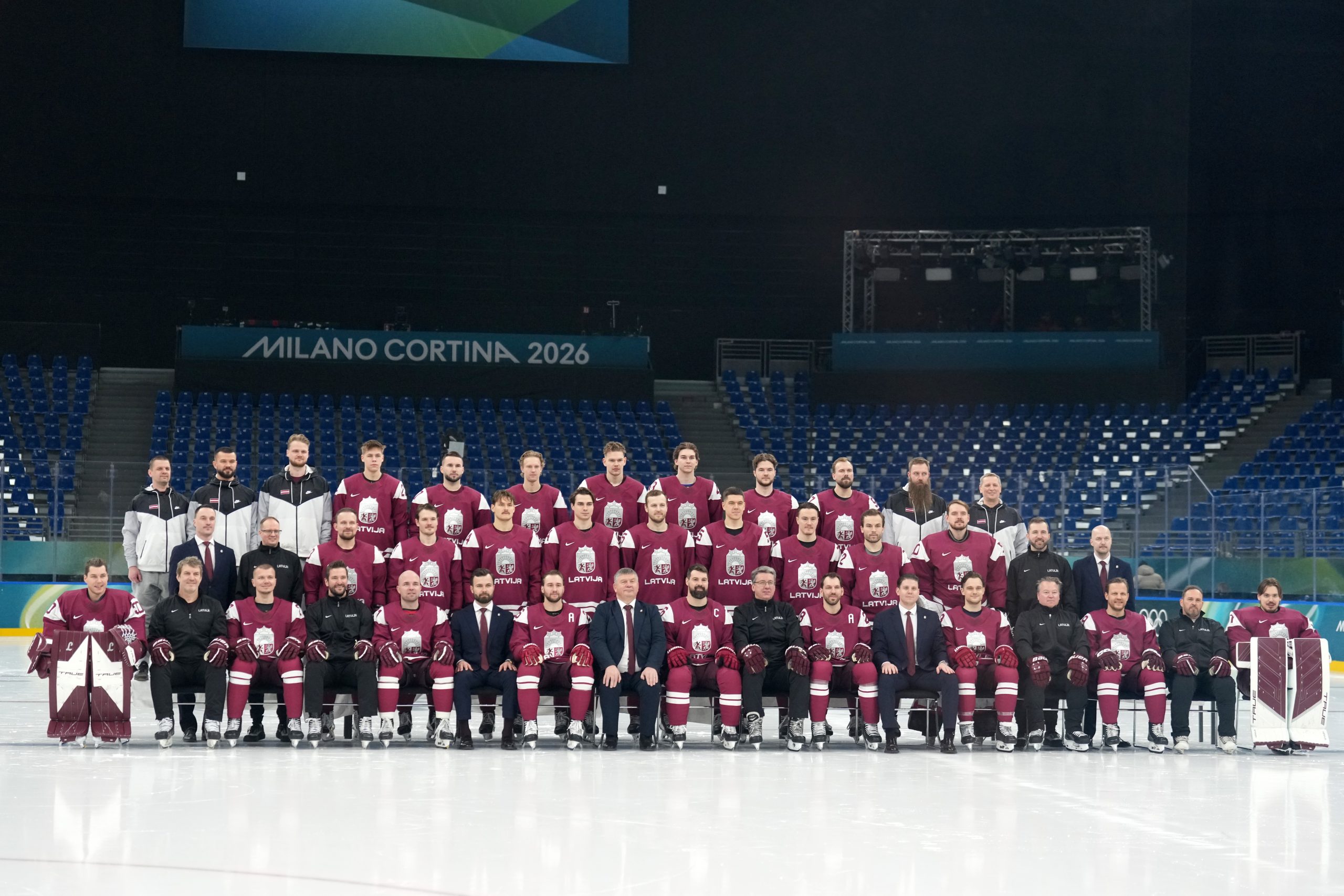 Latvijas hokeja izlase kopbildē uz ledus arēnā ar uzrakstu “Milano Cortina 2026”.