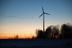 Vēja turbīna ziemas ainavā saulrieta laikā, redzami arī elektrolīniju balsti.