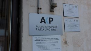 Uzņēmuma “Auces komunālie pakalpojumi” informatīvā plāksne pie ēkas.