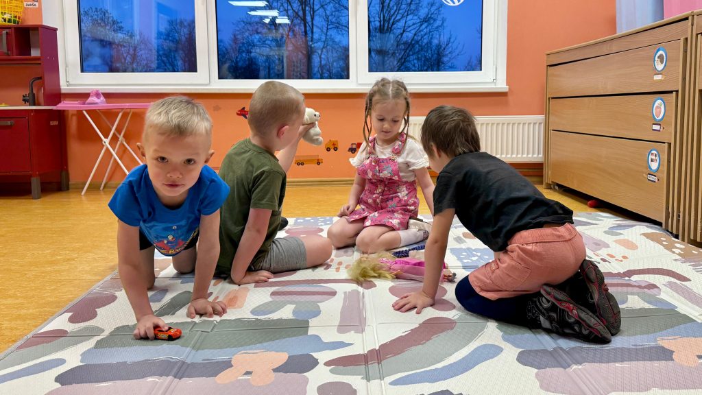 Četri bērni bērnudārzā rotaļājas uz krāsaina paklāja ar rotaļu automašīnām un lellēm.