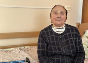 Seniore sēž istabā sociālās aprūpes centrā, blakus redzams spieķis