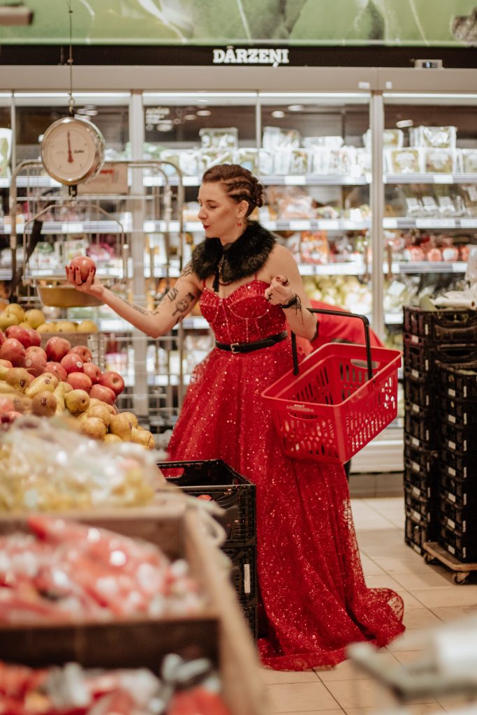 Evika Muizniece sarkanā balles kleitā izvēlas ābolus pārtikas veikalā.