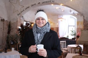 Diāna Druvaskalna Smēdes namā tur rokās baltu krūzi, fonā redzama vēsturiska interjera arka ar uzrakstu.
