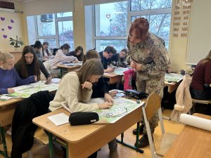 Skolotāja militārā formastērpā palīdz skolēniem analizēt kartes ģeogrāfijas stundā klasē.