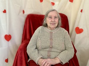 Seniore sēž krēslā uz sarkana fona ar sirsniņām Valentīndienas noformējumā