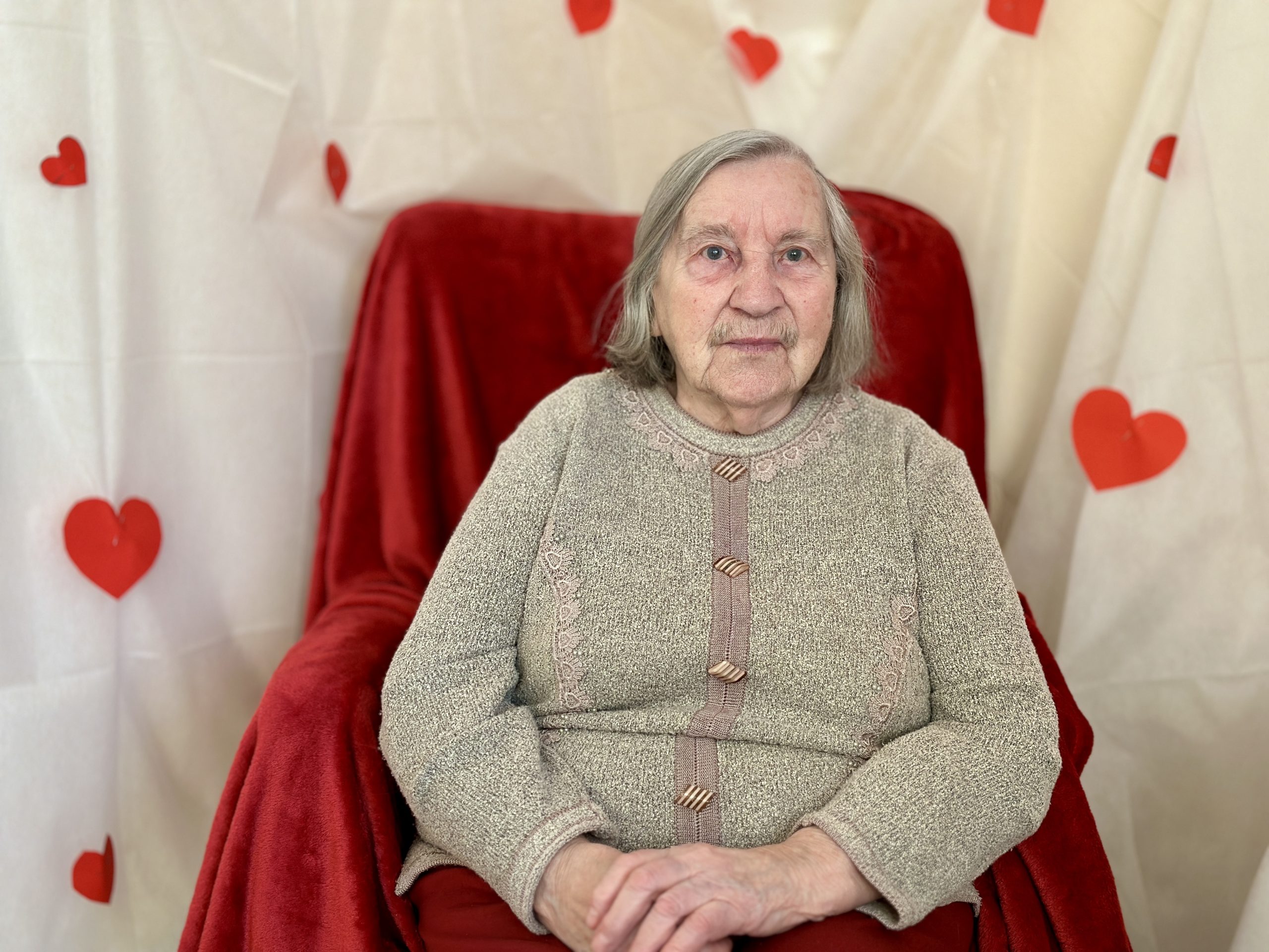 Seniore sēž krēslā uz sarkana fona ar sirsniņām Valentīndienas noformējumā