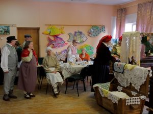 Cilvēki tautastērpos un tradicionālā apģērbā pulcējušies kultūras namā pie galda ar rokdarbiem un tējas galdu.