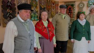 Cilvēki tradicionālā latviešu apģērbā kultūras namā, fonā redzami rokdarbu dekori un sienas paklāji.