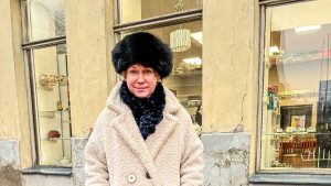 Kristīne Abika kažokādas cepurē un gaišā mētelī pie lietoto preču veikala skatloga