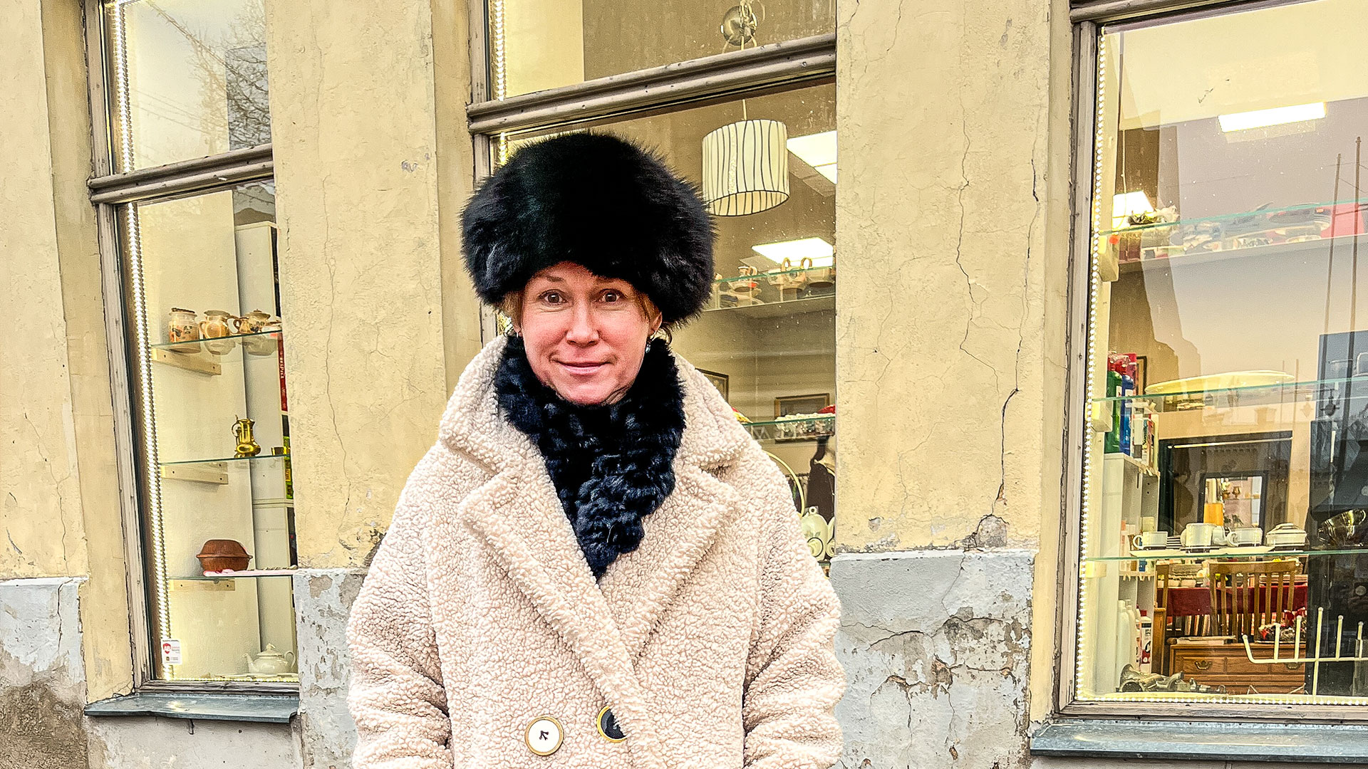 Kristīne Abika kažokādas cepurē un gaišā mētelī pie lietoto preču veikala skatloga