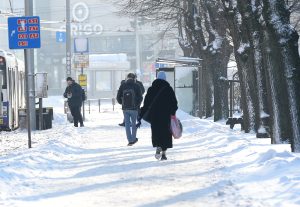 Gājēji iet pa sniegotu ietvi pie sabiedriskā transporta pieturas ziemas laikā