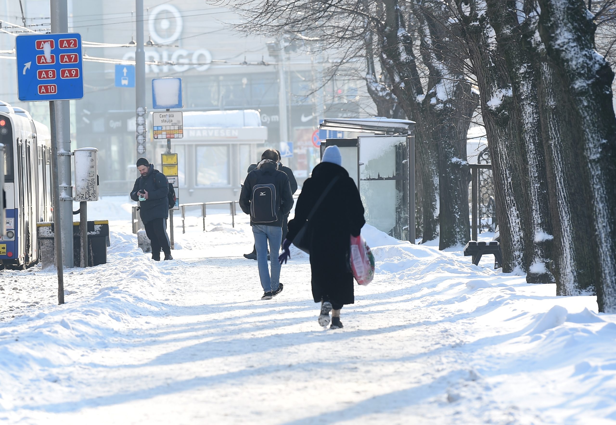 Gājēji iet pa sniegotu ietvi pie sabiedriskā transporta pieturas ziemas laikā