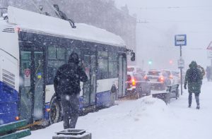 Sabiedriskais autobuss pieturā Rīgā spēcīga sniegputeņa laikā, fonā sastrēgums un gājēji sniegā.