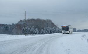 Autobuss brauc pa sniegotu lauku ceļu ziemas apstākļos, fonā mežs un snigšana.
