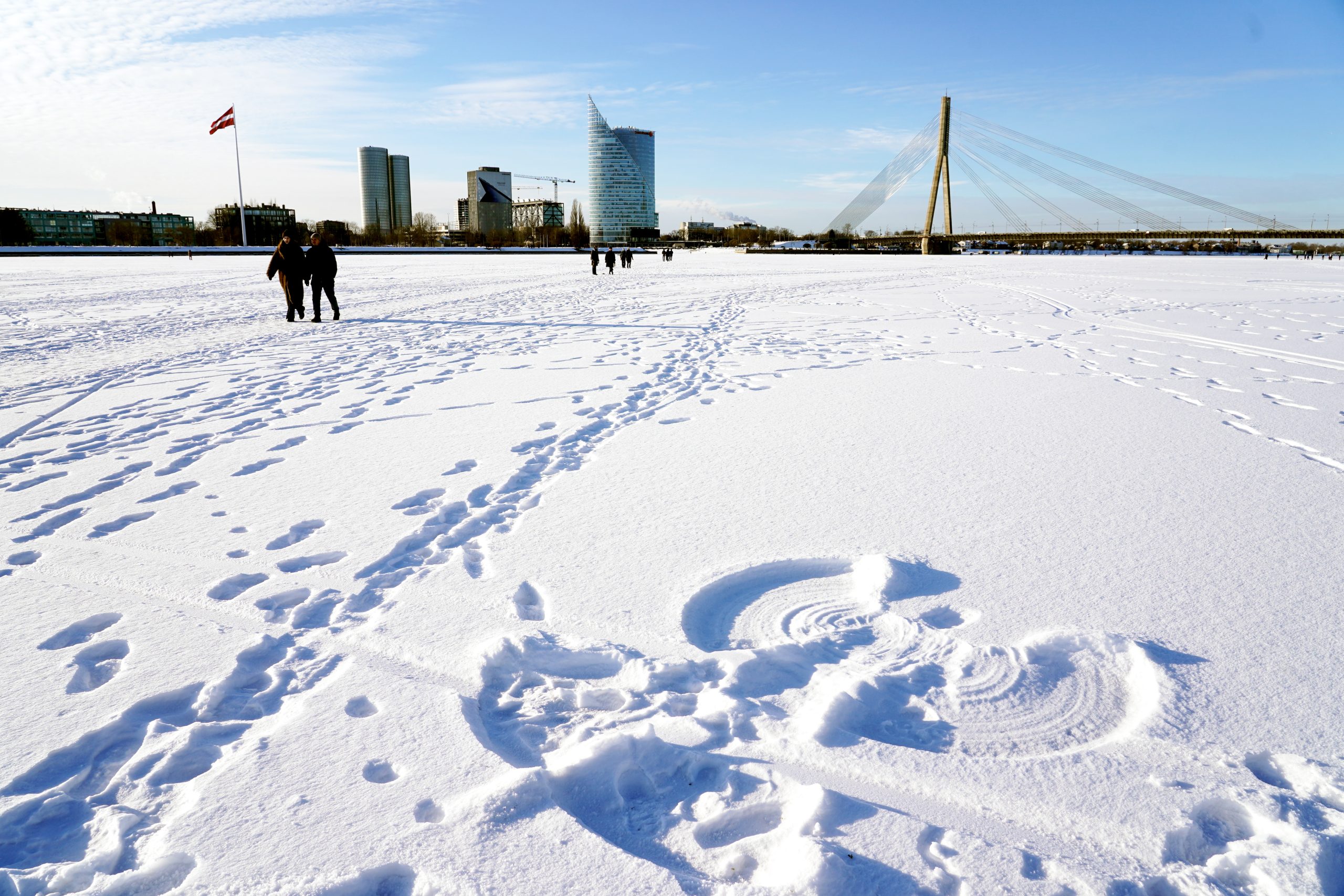 Cilvēki pārvietojas pa aizsalušo Daugavu Rīgas centrā ziemas laikā, fonā redzams Vanšu tilts un pilsētas panorāma.