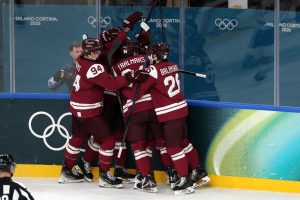 Latvijas hokeja izlases spēlētāji svin vārtu guvumu pie bortiem ar “Milano Cortina 2026” simboliku fonā.