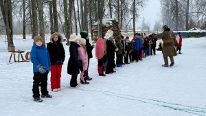 Bērni ziemas apģērbā stāv rindā sniegotā laukumā Meteņdienas pasākumā Leimaņos