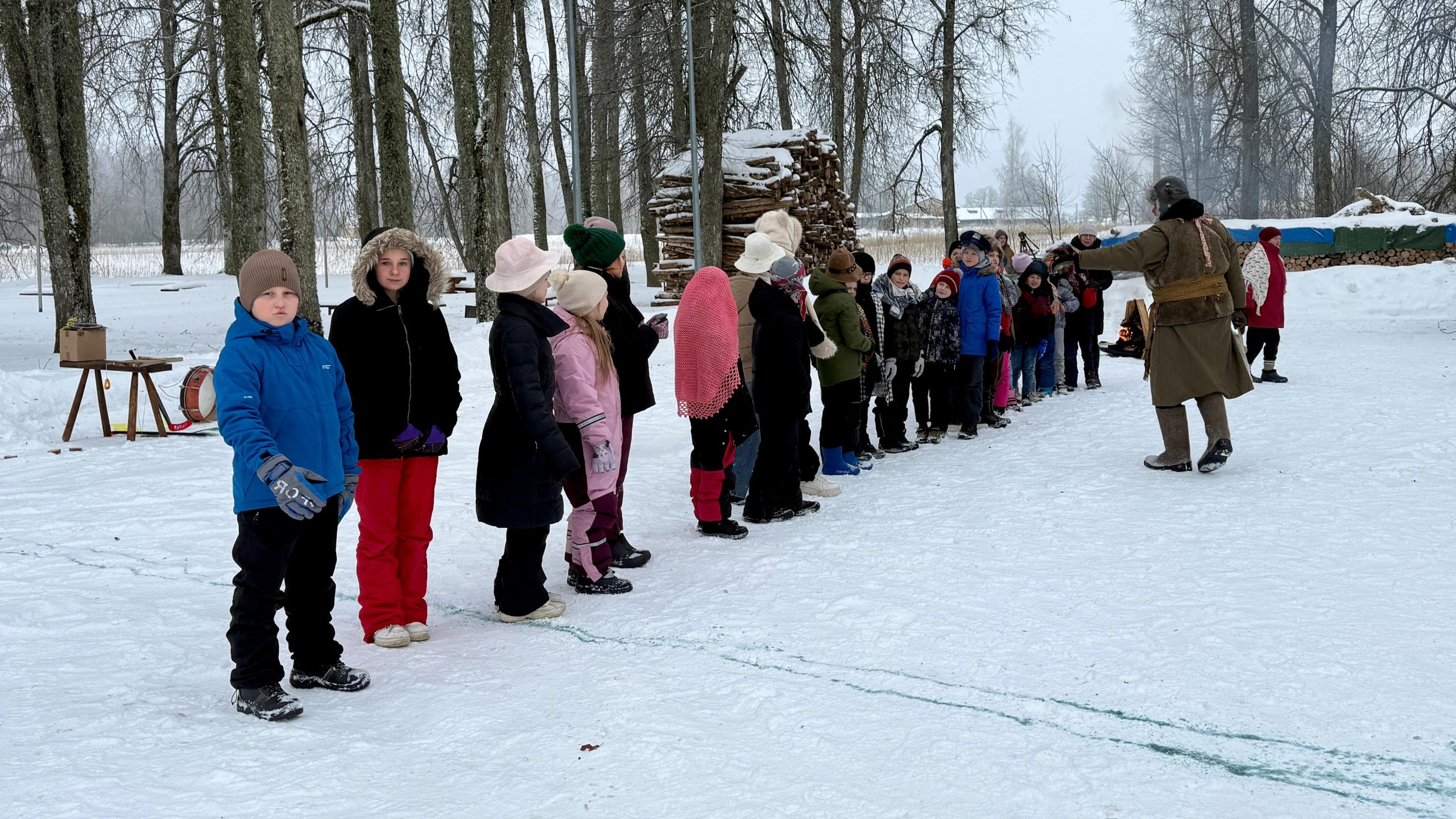 Bērni ziemas apģērbā stāv rindā sniegotā laukumā Meteņdienas pasākumā Leimaņos
