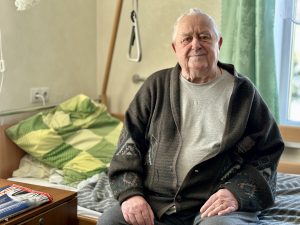 Sirmgalvis sēž uz gultas istabā pie loga, blakus redzams naktsskapītis ar žurnālu.