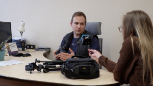 Žurnāliste ar mikrofonu intervē vīrieti birojā, uz galda redzama kamera un filmēšanas aprīkojums.