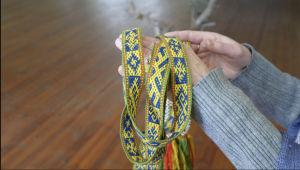 Rokās turēta krāsaina austā josta ar tradicionālu latviešu rakstu dzeltenā un zilā krāsā.