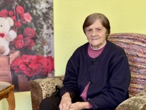 Sirmgalve sēž krēslā iekštelpās, fonā redzams ziedu plakāts ar sarkanām rozēm.