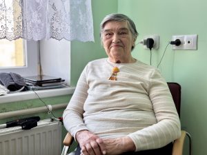 Sirmgalve sēž pie loga un smaida, rokām saliktām klēpī.