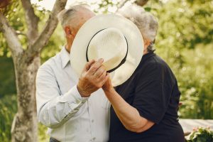 Vecāka gadagājuma pāris dārzā vasarā, vīrietis ar salmu cepuri aizsedz abu sejas.
