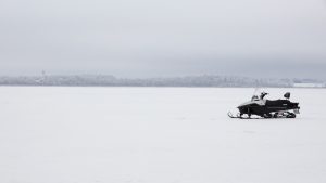 Sniega motocikls stāv uz aizsaluša ezera ziemas apstākļos ar miglainu ainavu fonā.