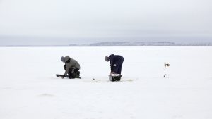 Divi cilvēki makšķerē uz aizsaluša ezera ziemā, izmantojot ledus urbjus un inventāru.
