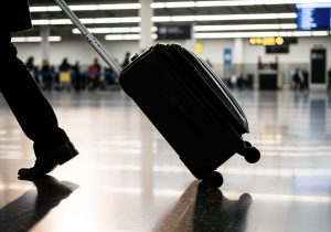 Cilvēks lidostā velk melnu ceļasomu uz riteņiem, fonā redzama izplūdusi pasažieru zona un izlidošanas tablo.