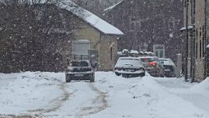 Spēcīga snigšana pilsētas ielā, automašīnas pārvietojas pa sniegotu un grūti izbraucamu ceļu.