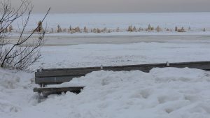 Apsnigusi koka soliņa pie jūras ziemā, fonā aizsalusi piekraste un ledus klāta ūdens virsma.