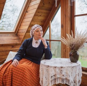 Sieviete tradicionālā latviešu apģērbā sēž pie galda ar mežģīņu galdautu koka interjerā pie loga.