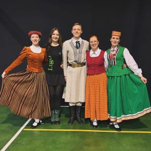 Grupa jauniešu un dejotāju latviešu tautastērpos pozē kopīgai fotogrāfijai uz skatuves.