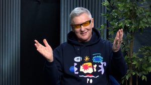 Roberts Gobziņš tumšā džemperī ar krāsainu apdruku un dzeltenām brillēm studijā
