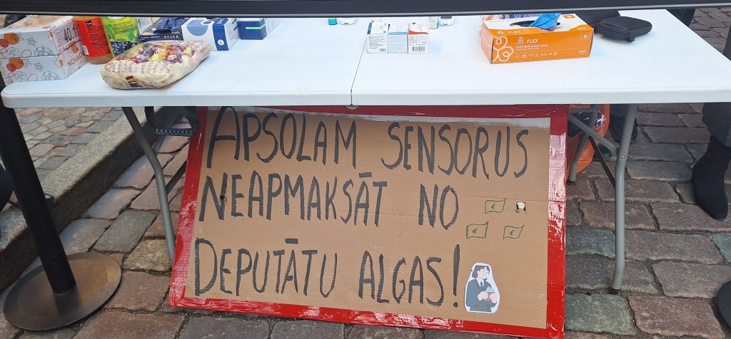 Plakāts ar uzrakstu par sensoru neapmaksāšanu deputātu algu vietā