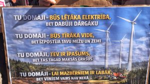 Protesta plakāts pret vēja parkiem ar tekstu par elektrības cenām un vidi.