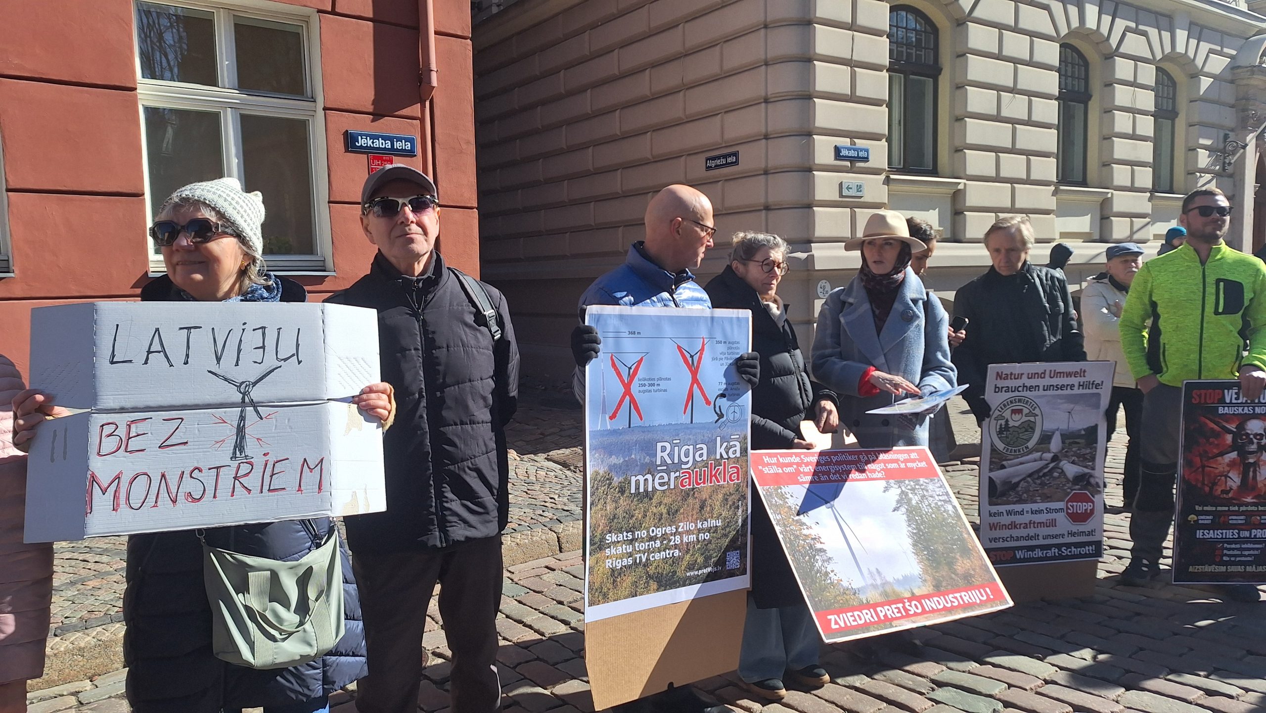 Cilvēki ar plakātiem piedalās protestā pret vēja parkiem pilsētvidē.