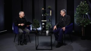 Atis Zviedris un Aivars Lapšāns sarunājas tumšā studijā, sēžot krēslos pie neliela galdiņa
