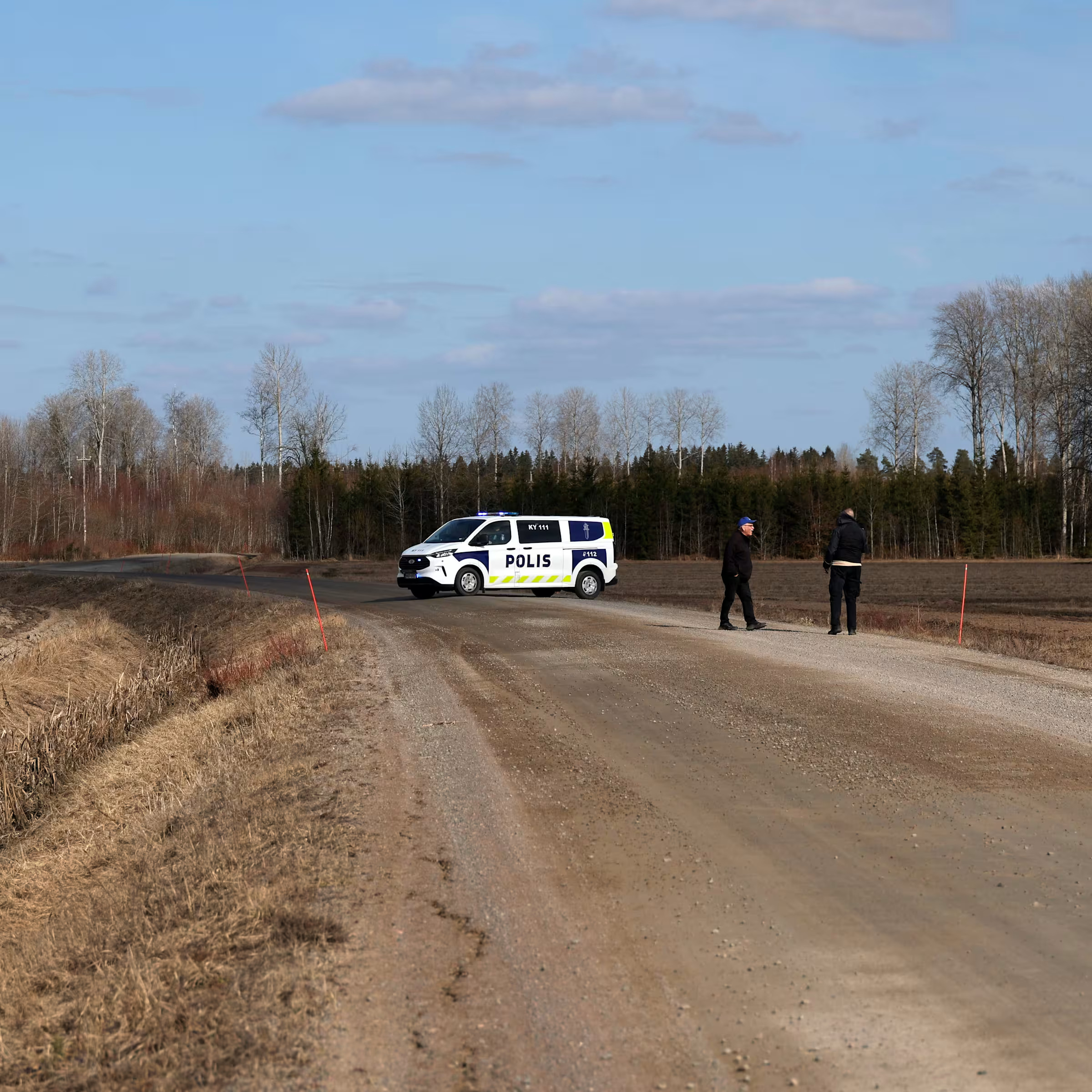 Policijas automašīna bloķē ceļu lauku teritorijā, divi cilvēki stāv blakus