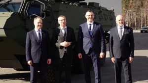 Vīrieši uzvalkos stāv pie militārā bruņutransportiera rūpnīcas teritorijā