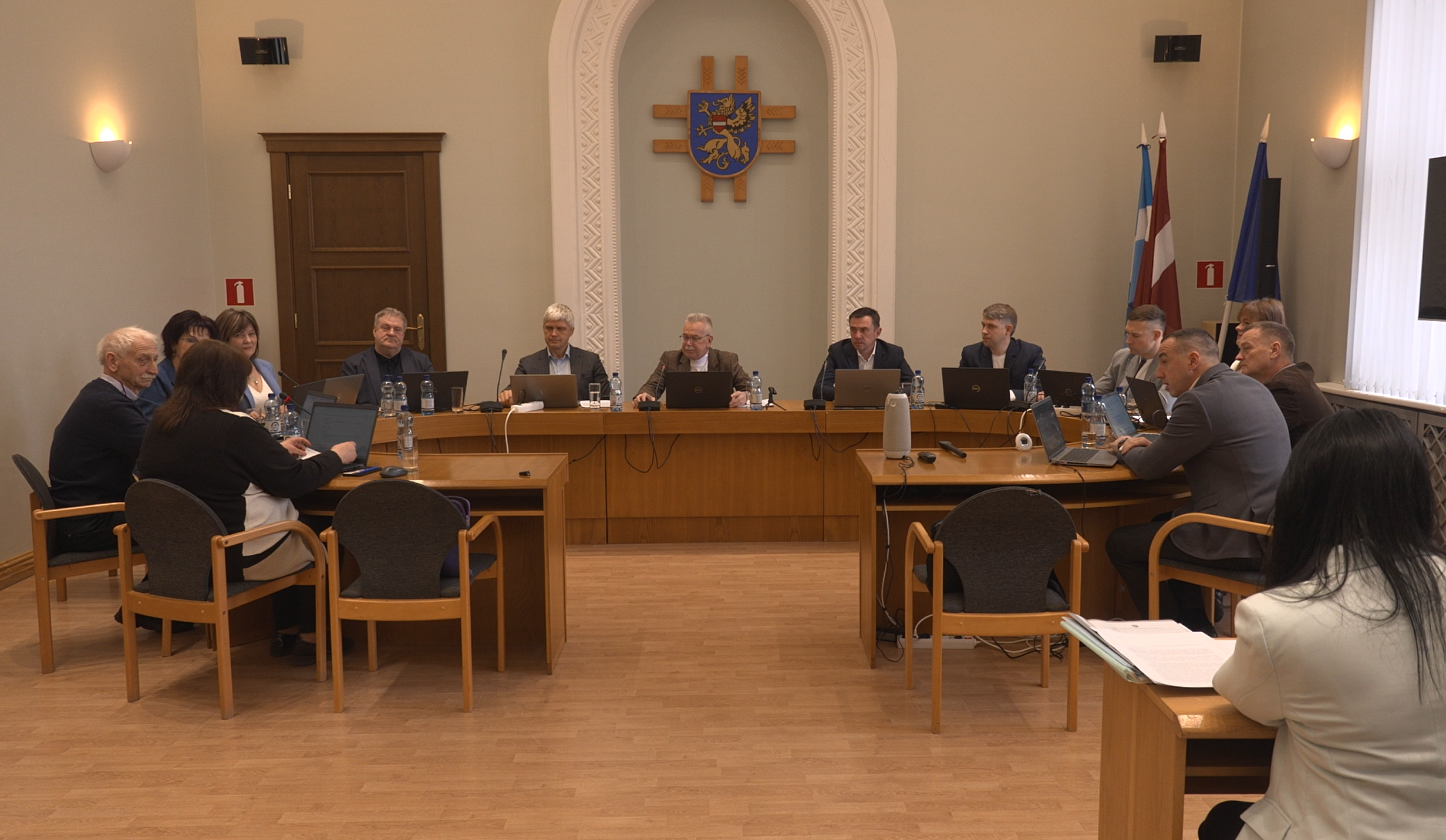 Pašvaldības sēde ar deputātiem pie galda un Latvijas karogiem fonā