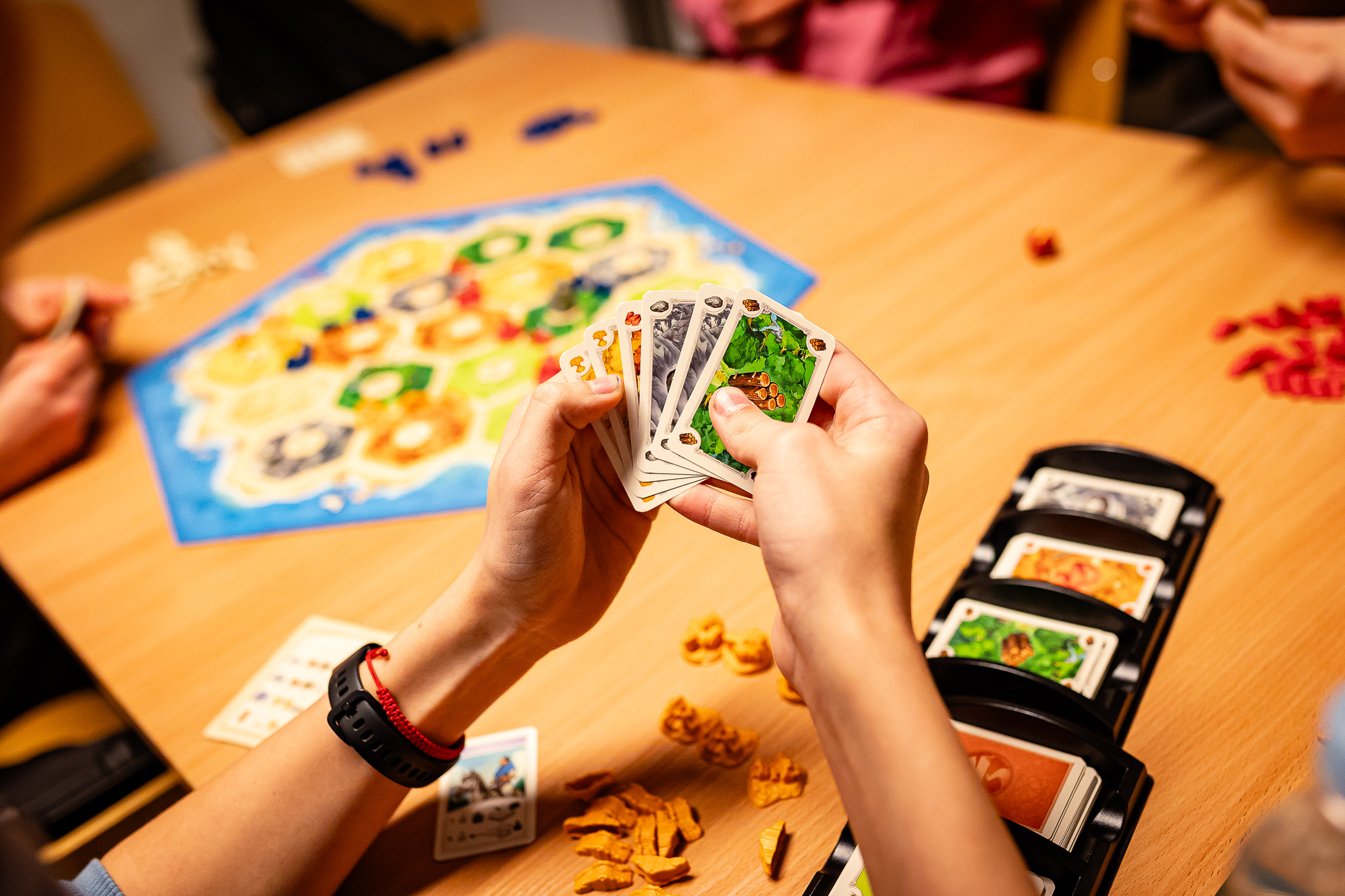 Spēlētāji pie galda spēlē stratēģijas galda spēli „CATAN“, rokās turot resursu kārtis pie spēles galda.