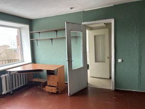 Istaba sociālajā dzīvoklī ar rakstāmgaldu un plauktiem pie sienas