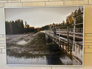Fotogrāfiju izstādē redzams attēls ar cilvēkiem uz tilta pār upi rudenīgā ainavā.
