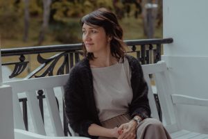 Sieviete sēž uz baltas soliņa ārā un raugās sānis rudens ainavā