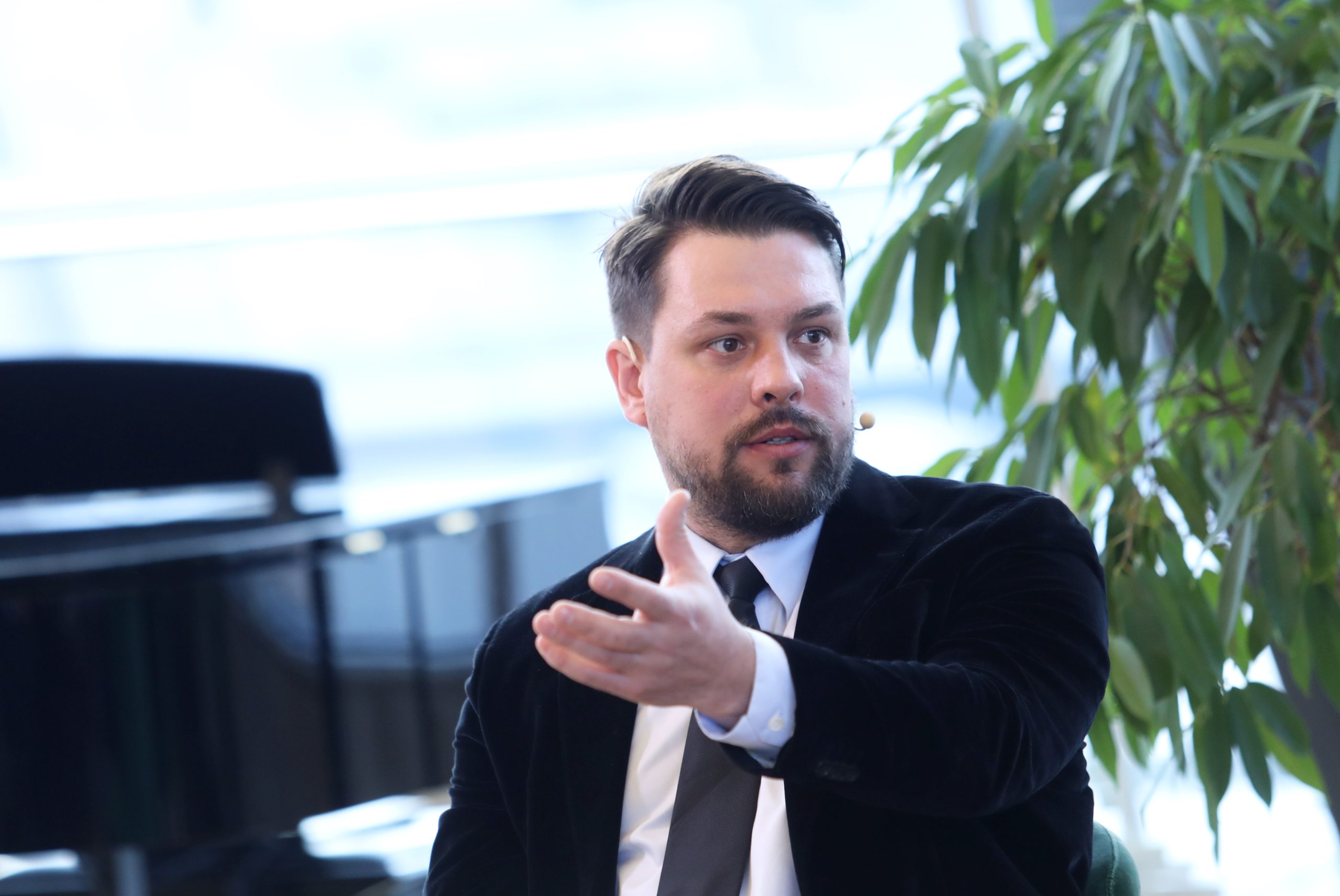 Sandis Šrāders runā pasākumā ar mikrofonu un žestikulē ar roku